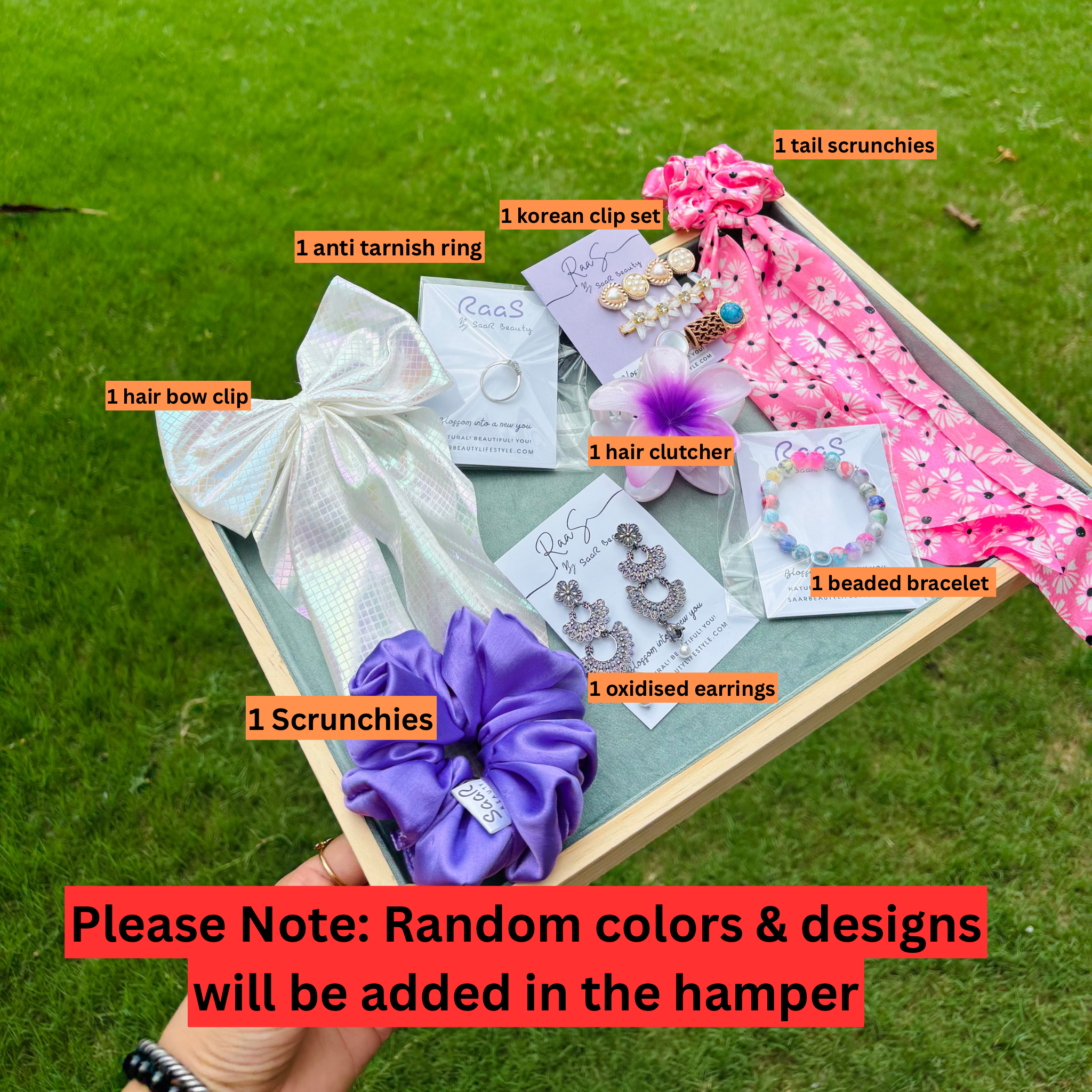 Box of jewelry and hair accessories with labels on a grass background Premium Rakshabandhan gift box with satin scrunchie, bow clip, jhumka earrings, flower claw, bracelet, ring, tail scrunchie, and Korean clip set by Saar Beauty