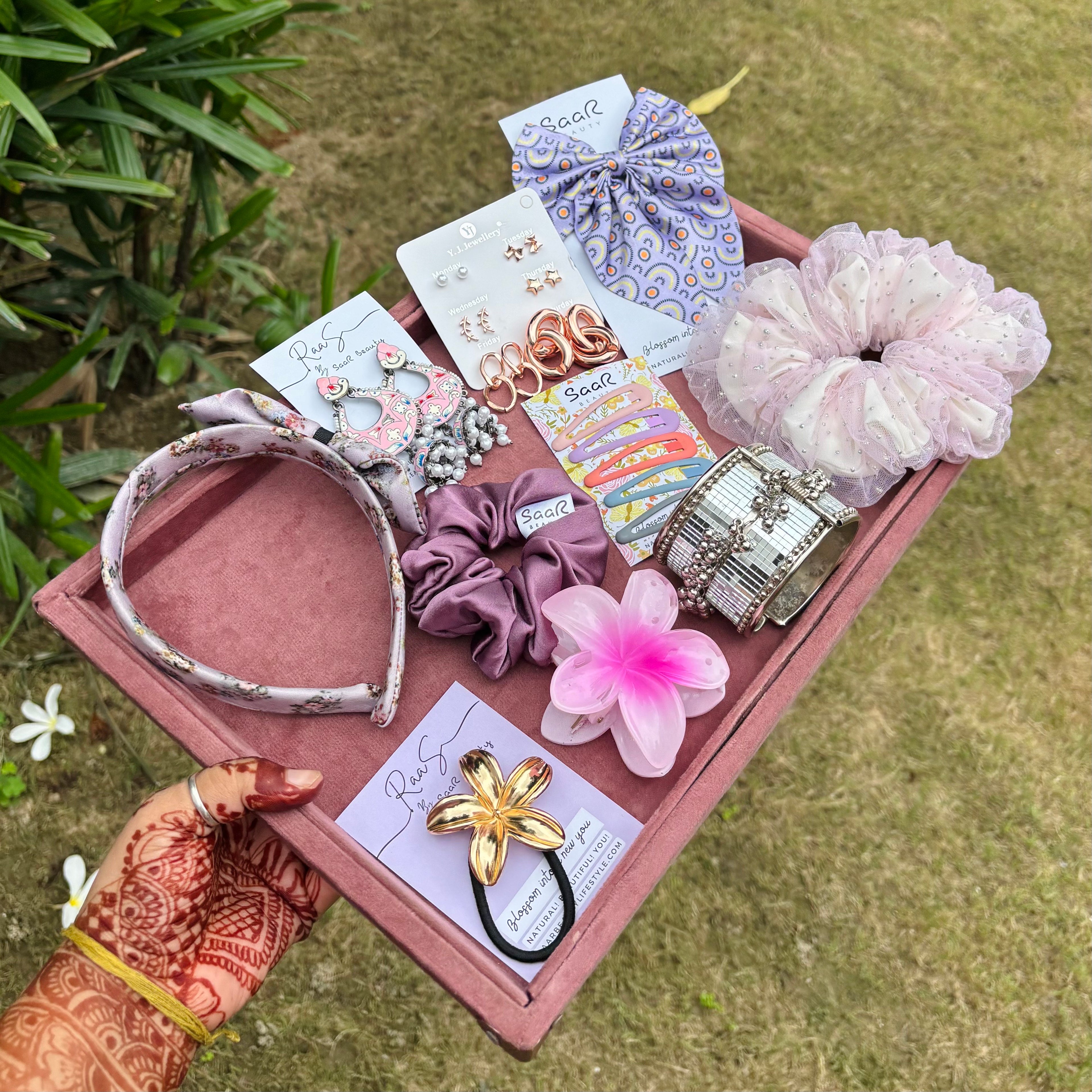 Collection of hair accessories including headbands, scrunchies, and clips on a pink tray outdoors.Rakshabandhan Gift Box with jhumka earrings, satin scrunchies, fluffy bow, floral claw clip, satin headband, kada bracelet, and tic tac clips by Saar Beauty.