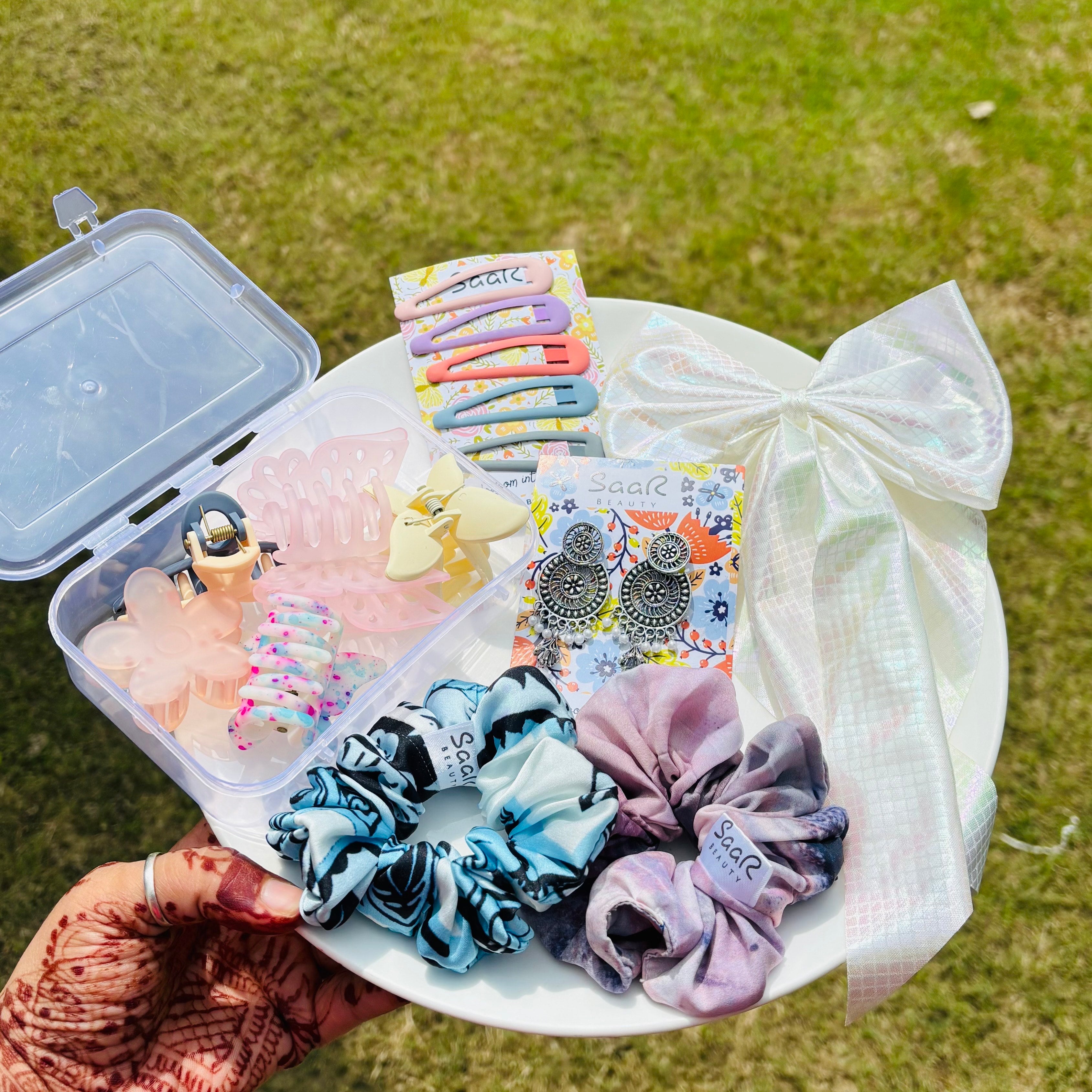 Assorted hair accessories including scrunchies, clips, and earrings on a white plate outdoors. Premium Rakhi gift set by Saar Beauty with hair bow, jhumka earrings, tic tac clips, two printed scrunchies, and five hair claws in a clear plastic box