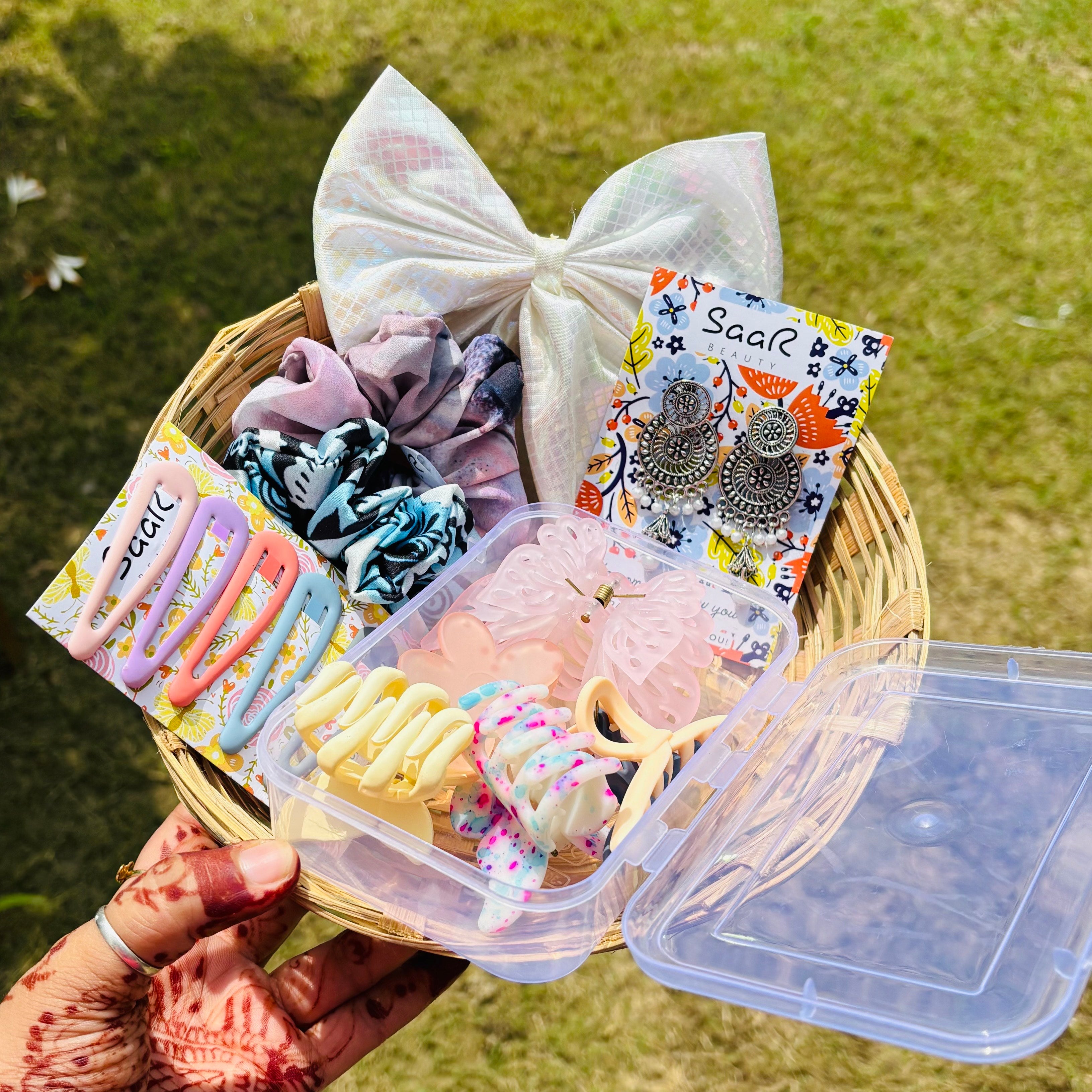 Basket with hair accessories including bows, clips, and pins on a grassy background Premium Rakhi gift set by Saar Beauty with hair bow, jhumka earrings, tic tac clips, two printed scrunchies, and five hair claws in a clear plastic box