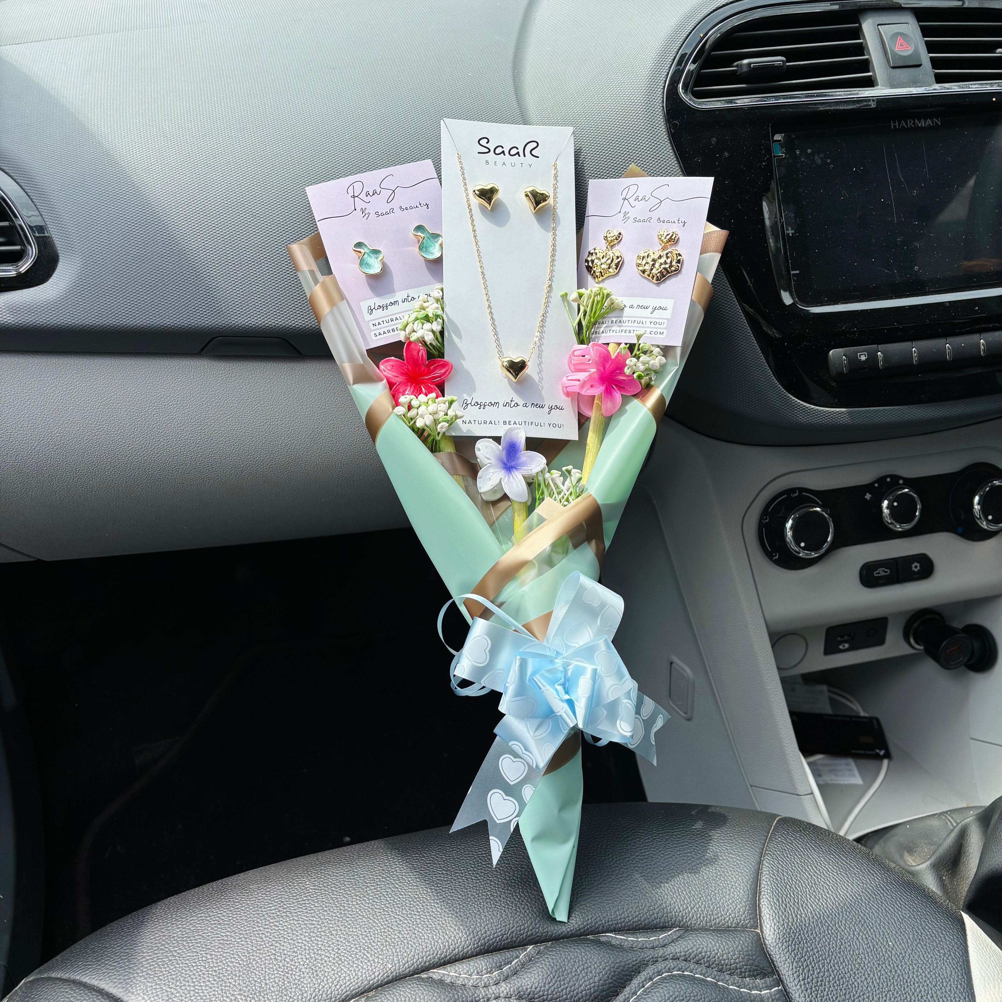 Bouquet of flowers with jewelry cards on a car dashboard Premium gift bouquet by Saar Beauty with anti-tarnish heart necklace and earrings, pastel floral wrapping, and small flower claw hair clips. Ideal for thick hair, long hair, and gifting special women and girls.