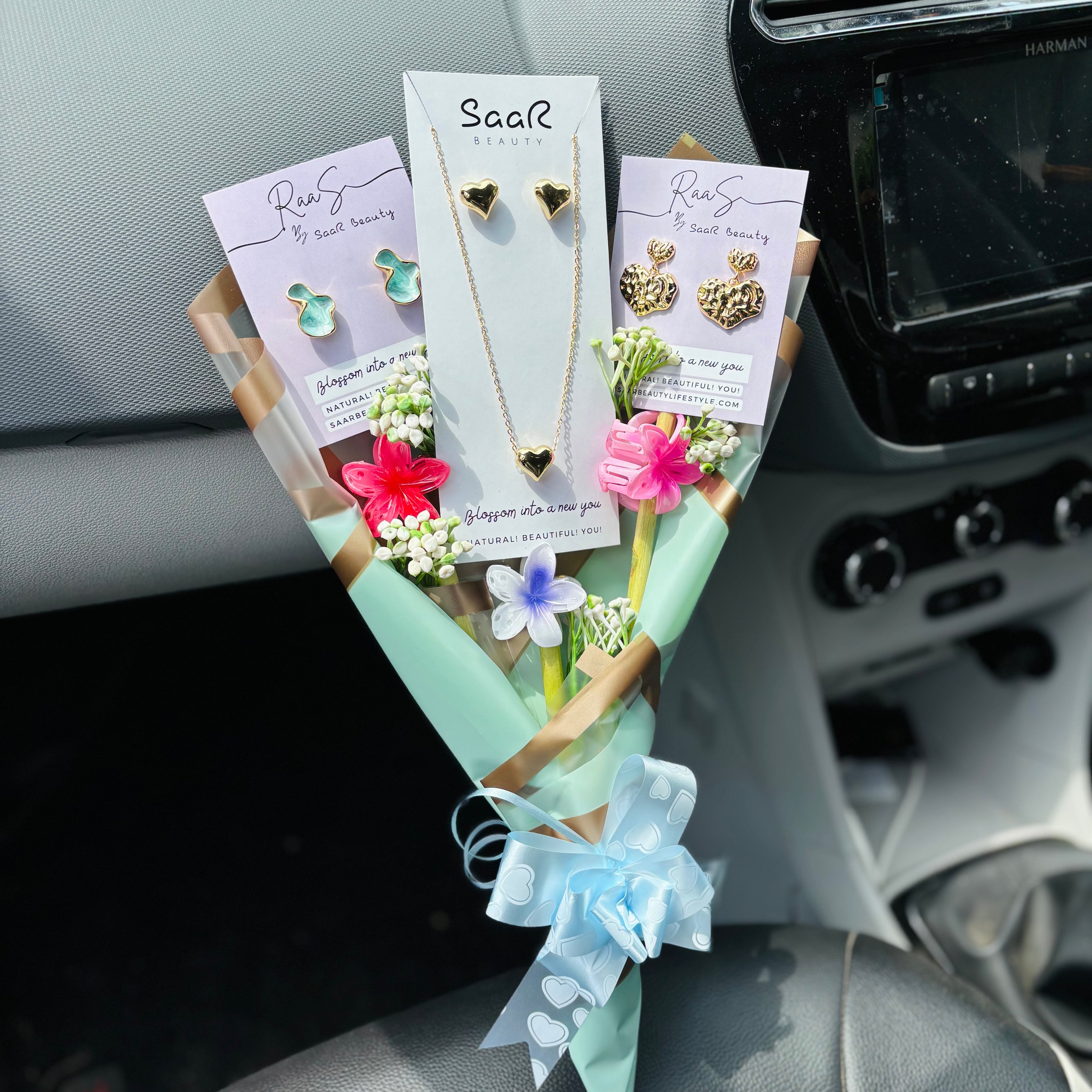 Jewelry set with necklaces and earrings displayed on a car dashboard.Premium gift bouquet by Saar Beauty with anti-tarnish heart necklace and earrings, pastel floral wrapping, and small flower claw hair clips. Ideal for thick hair, long hair, and gifting special women and girls.