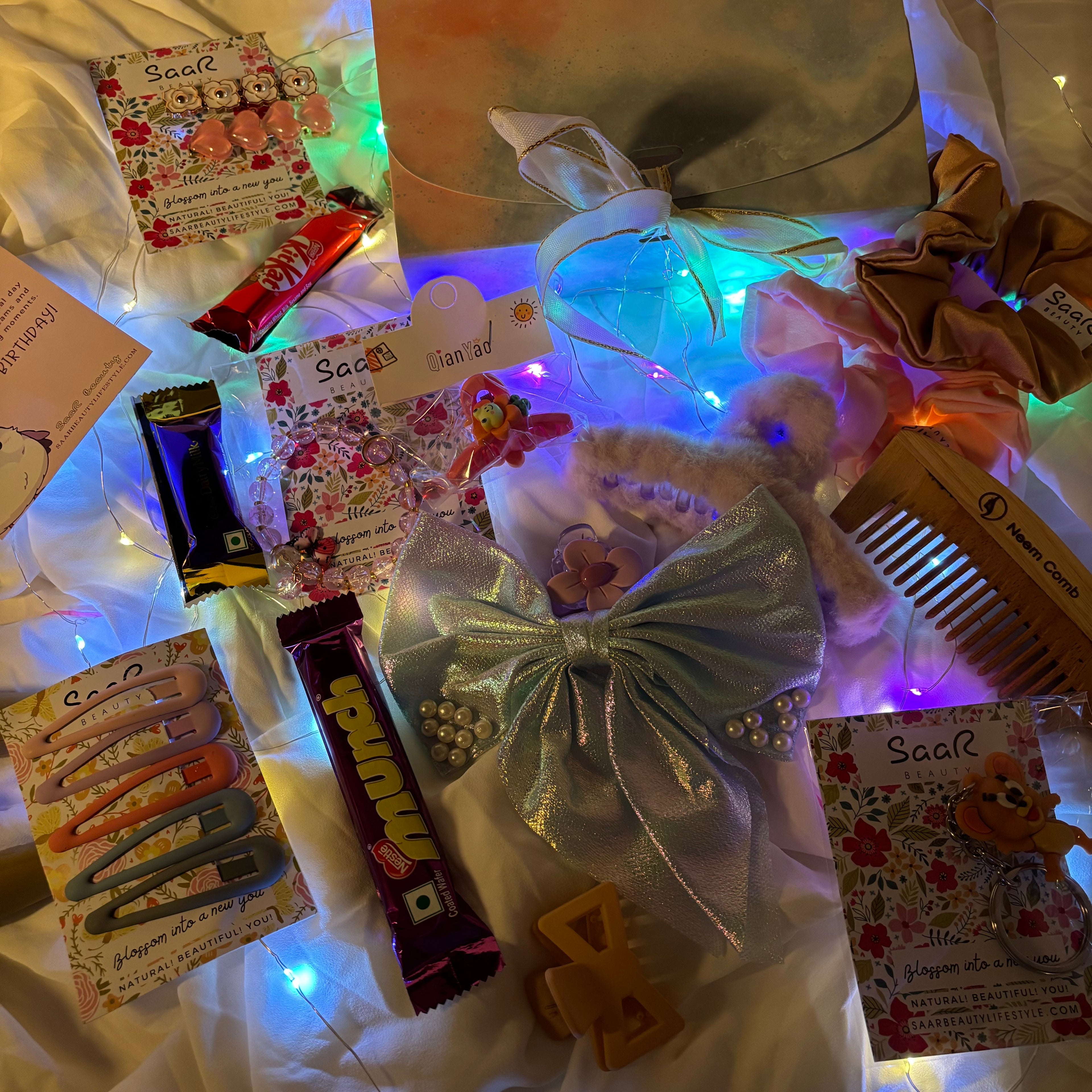 Assorted items including a teddy bear, hair accessories, and candy on a white surface with colorful lights."Luxury birthday surprise gift box for girls with chocolates and accessories"