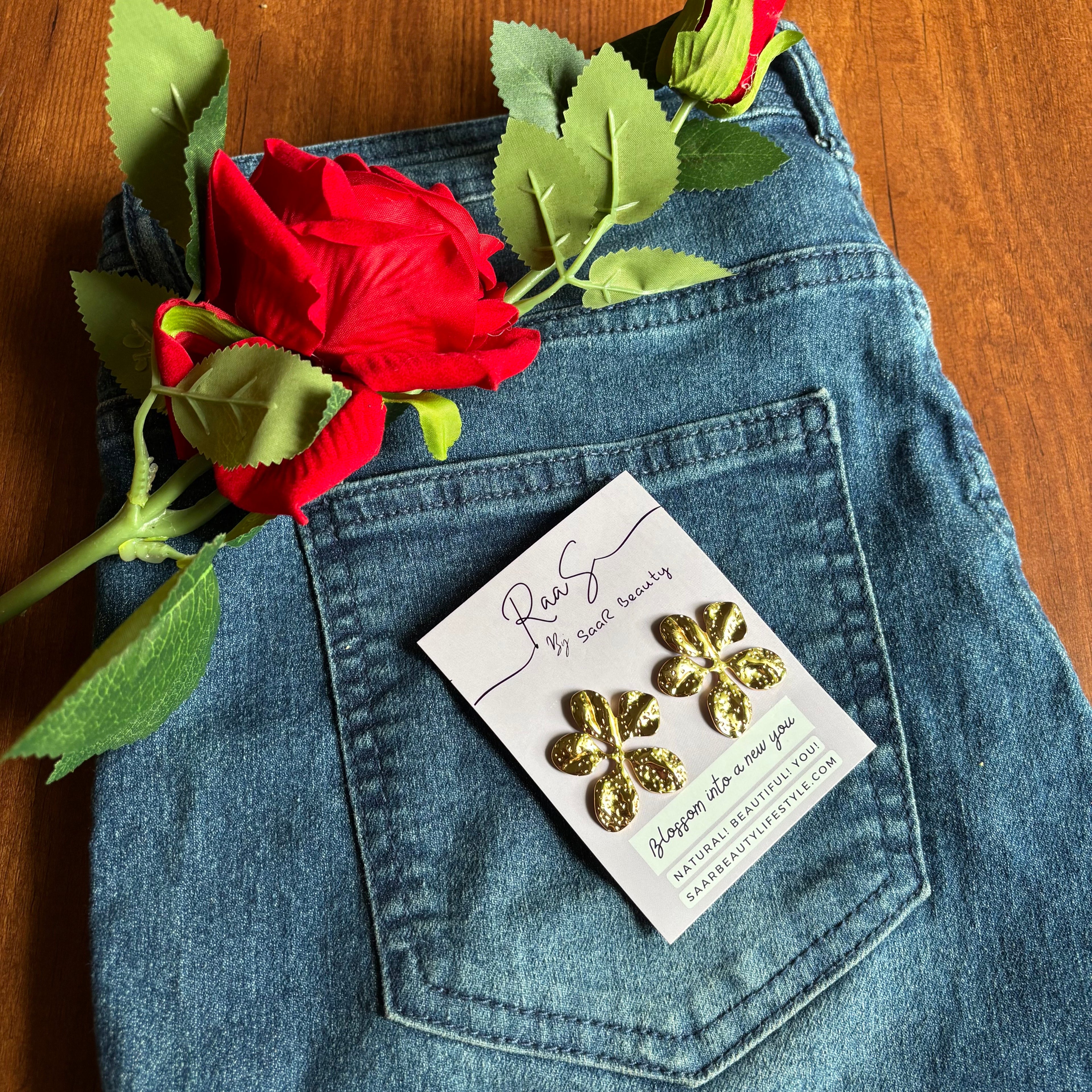Gold floral earrings on a card in a pocket of blue jeans with a red rose and green leaves. Elegant flower stud earrings โ anti-tarnish jewelry by Saar Beauty
