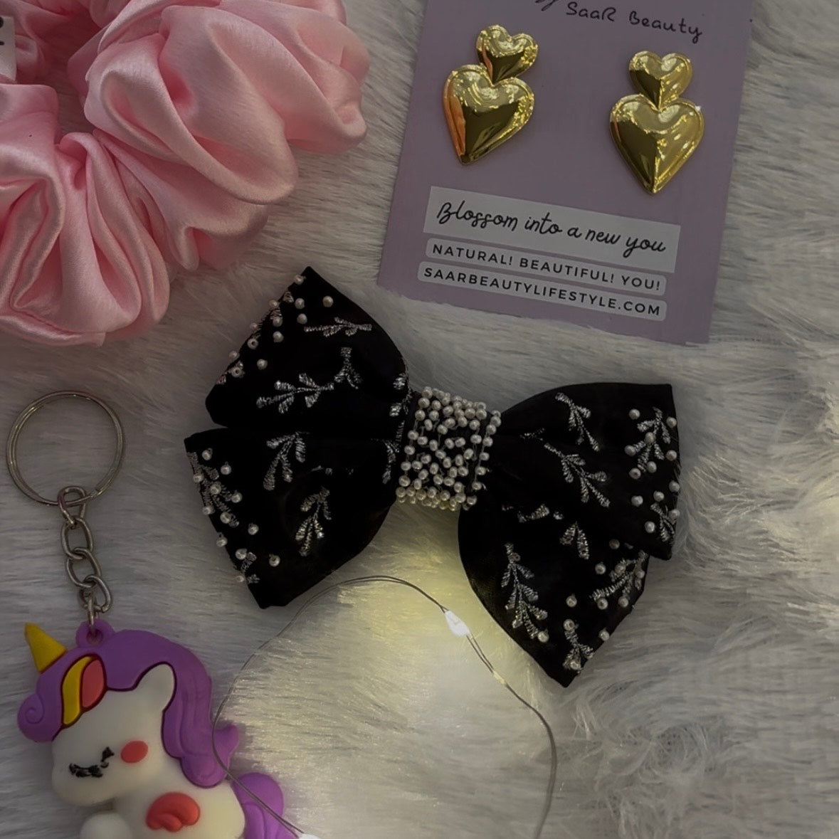 Black bow with white details, gold heart-shaped earrings, pink scrunchies, and a unicorn keychain on a textured surface.
