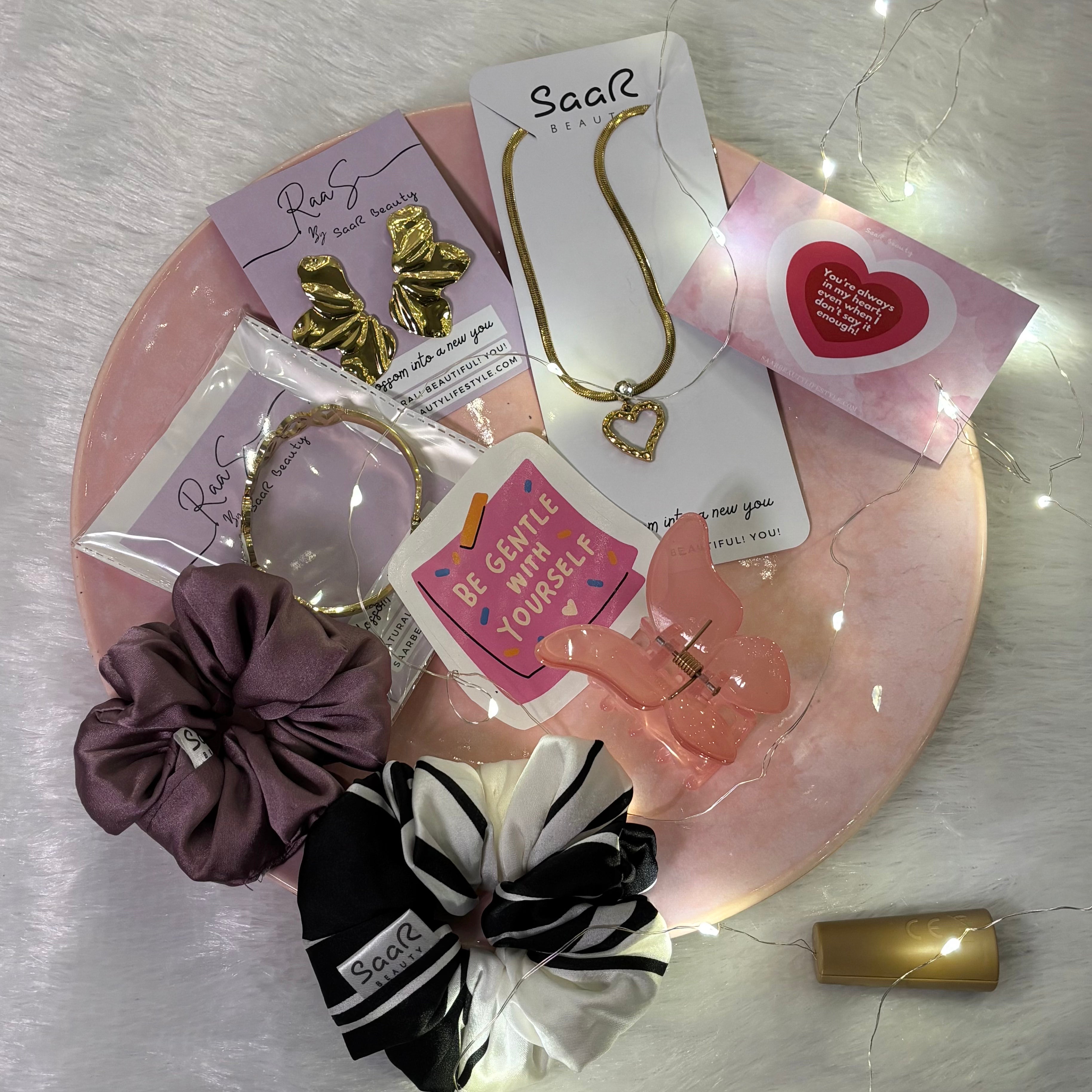 Collection of hair accessories and jewelry on a pink plate with decorative lights. Saar Beauty Buy 3 Get 5 Free Hamper – Includes waterproof jewelry, scrunchies, butterfly clip, fairy lights, and gifting card.