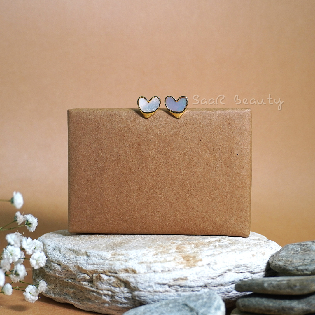 Close-up of lightweight heart earrings in white enamel with gold accents, styled on a neutral backdrop.