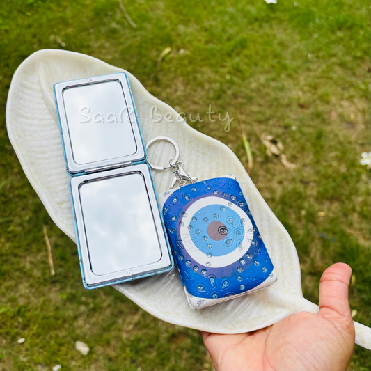 Compact mirror and blue evil eye keychain on a white cloth with grass  Cute Kitty and Evil Eye Keychain Pocket Mirrors – Portable & Stylish Design by Saar Beautybackground