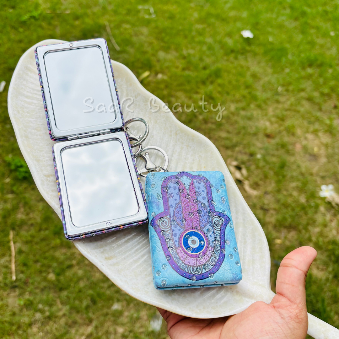 Blue and purple hamsa keychain with mirror on a white leaf against a green grass background Cute Kitty and Evil Eye Keychain Pocket Mirrors – Portable & Stylish Design by Saar Beauty