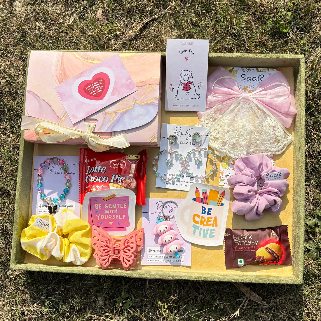 Gift box with stationery items, snacks, and decorative elements on a grassy Saar Beauty luxury light gift hamper for girls with accessories and chocolates
Premium light gift hamper for women with jewellery and cute accessories background 