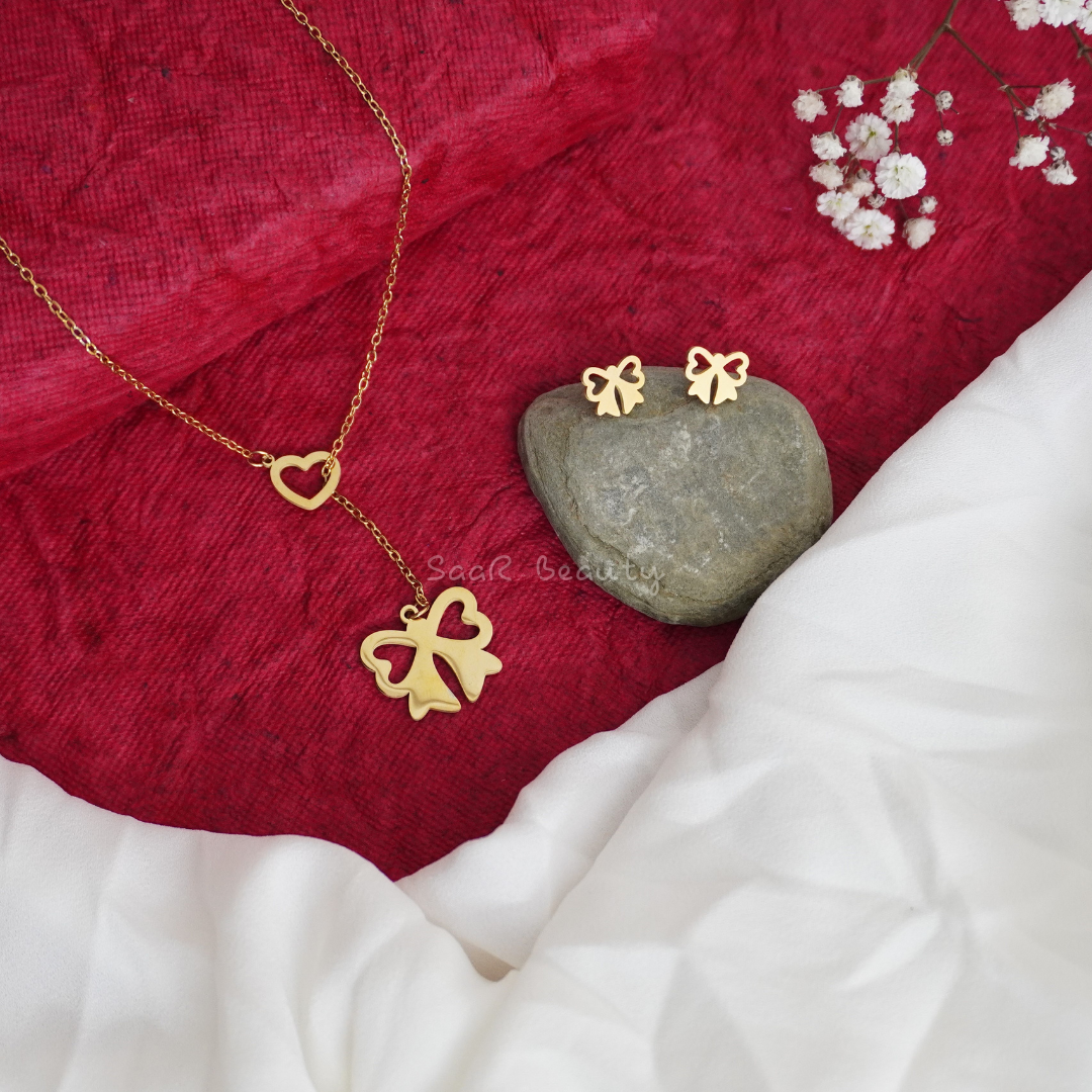 Gold-plated bow necklace and heart charm with matching stud earrings on a red fabric background.