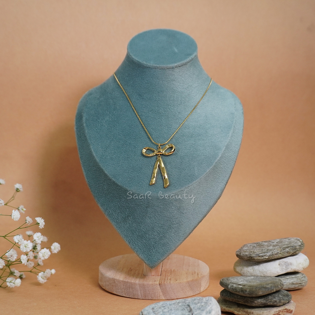 Gold-tone bow-shaped stainless steel pendant necklace displayed on a blue velvet stand, styled with delicate flowers and natural stones.