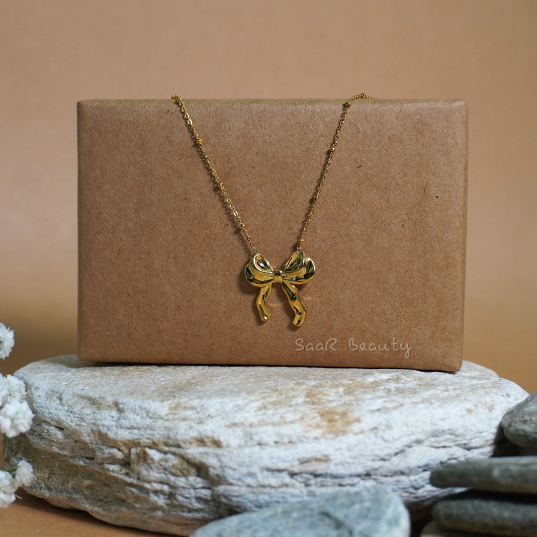 Golden bow charm necklace displayed elegantly on a brown box with minimalist styling by SaaR Beauty.