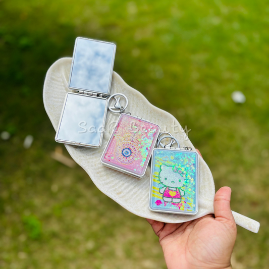 Three small compact mirrors with colorful designs held by a hand against a grassy background. Portable travel mirror keychain with glitter design