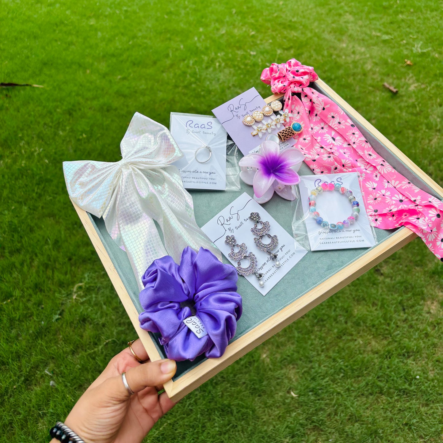 Gift box with hair accessories and jewelry on a grassy background Premium Rakshabandhan gift box with satin scrunchie, bow clip, jhumka earrings, flower claw, bracelet, ring, tail scrunchie, and Korean clip set by Saar Beauty