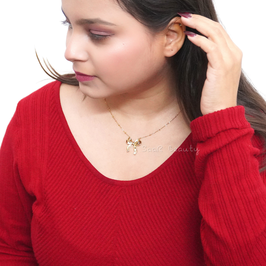 Model wearing SaaR Beauty's golden bow pendant necklace, paired with a red sweater, showcasing minimalist charm.