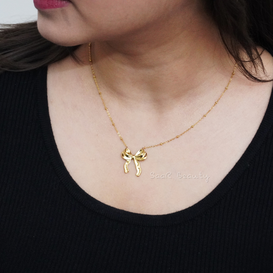 Model wearing SaaR Beauty's golden bow pendant necklace, paired with a red sweater, showcasing minimalist charm.