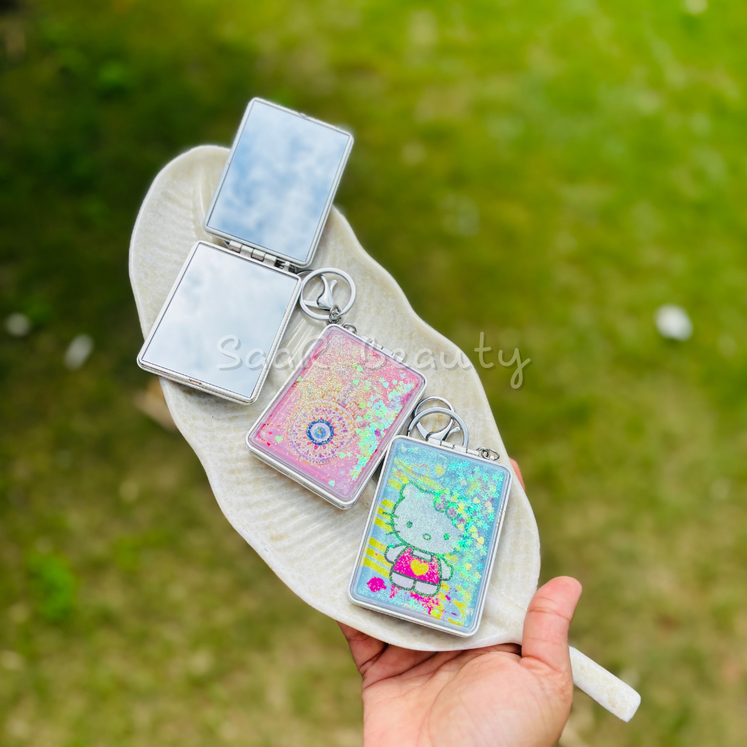 Three small rectangular keychains with colorful designs on a stone surface. Portable travel mirror keychain with glitter design