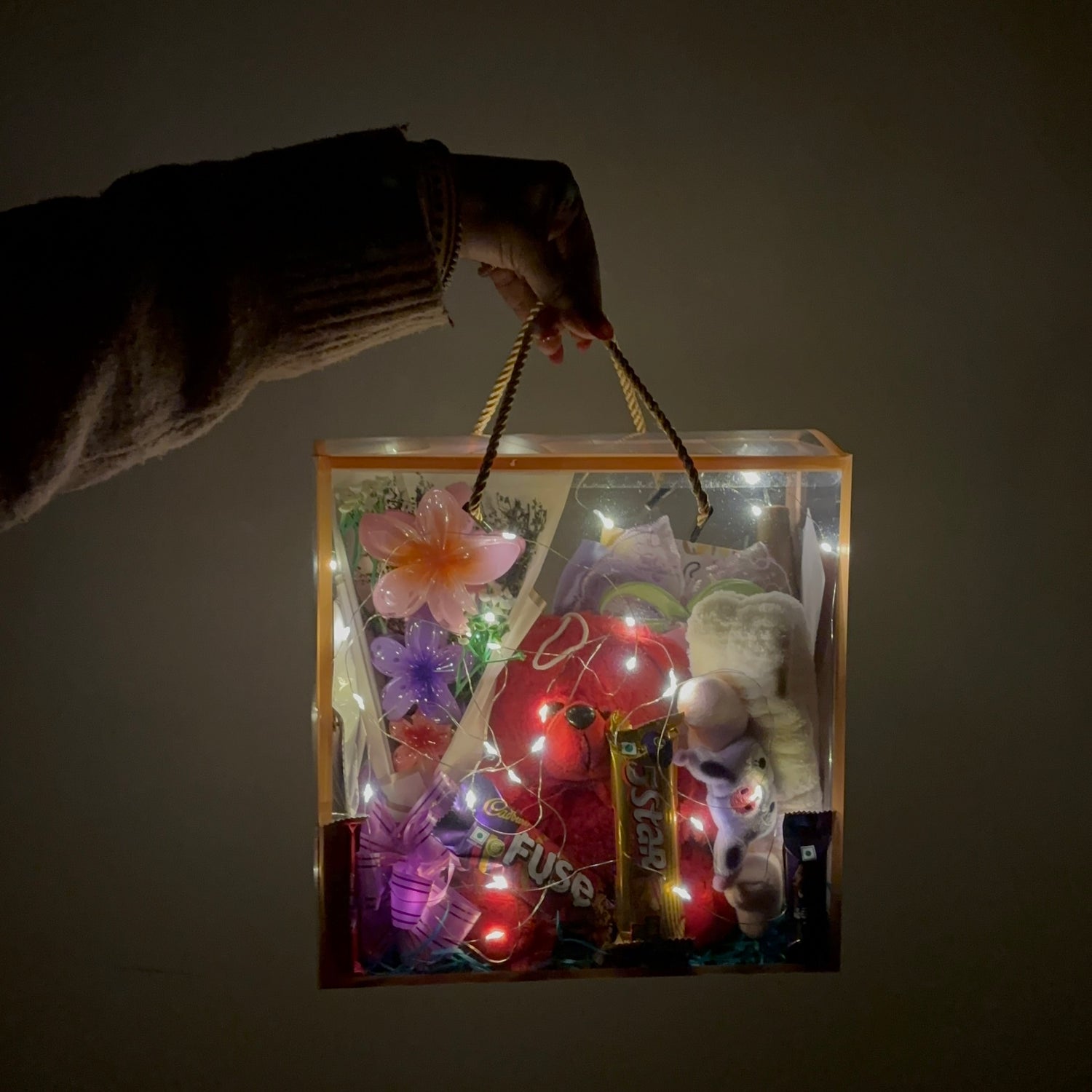 Gift bag with LED lights and a hand holding it against a dark background Premium gift box for women with beauty accessories teddy and chocolates