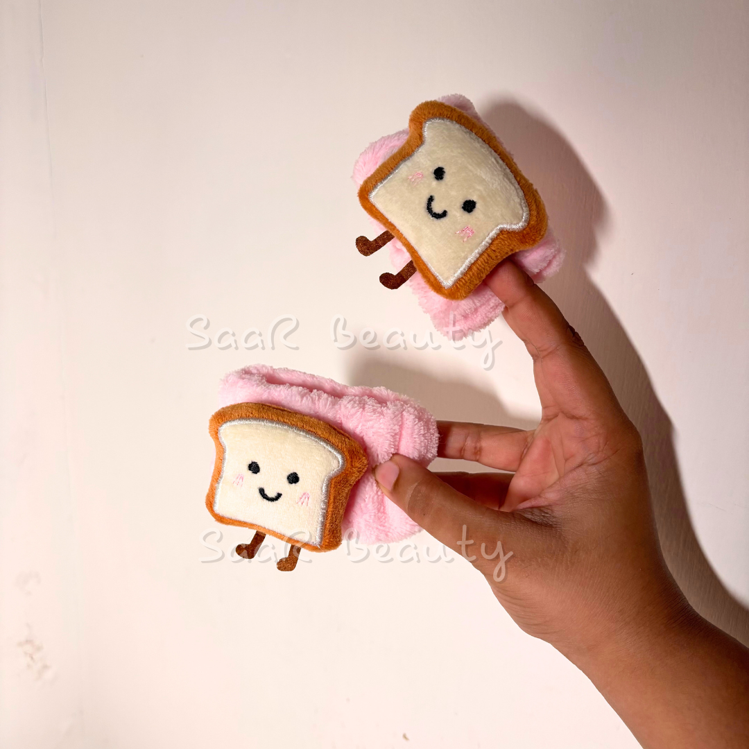 Hand holding two plush toys shaped like slices of bread with smiling faces on a light background. Pink plush skincare headband with bread design and water-absorbing hand wraps