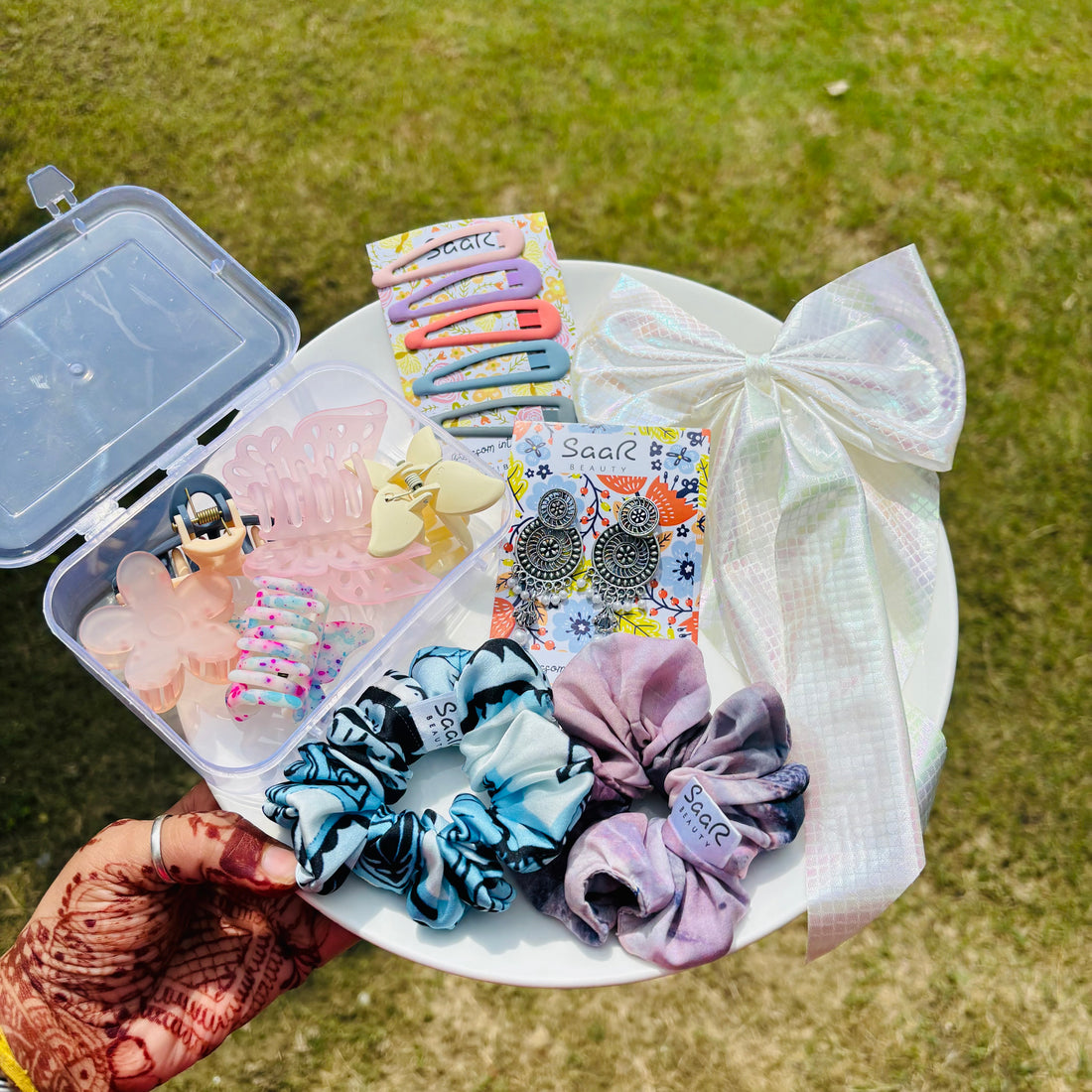 Assorted hair accessories including clips, scrunchies, and earrings on a white plate outdoors. Premium Rakhi gift set by Saar Beauty with hair bow, jhumka earrings, tic tac clips, two printed scrunchies, and five hair claws in a clear plastic box