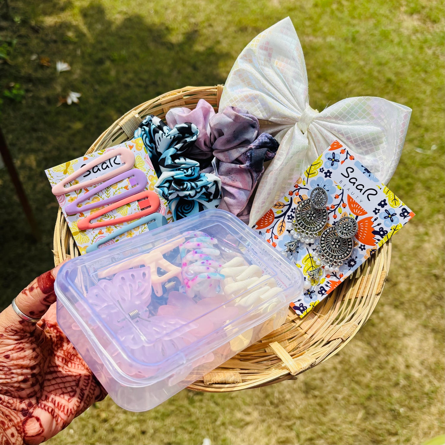 Basket with hair accessories including scrunchies, clips, and earrings on a grassy background Premium Rakhi gift set by Saar Beauty with hair bow, jhumka earrings, tic tac clips, two printed scrunchies, and five hair claws in a clear plastic box