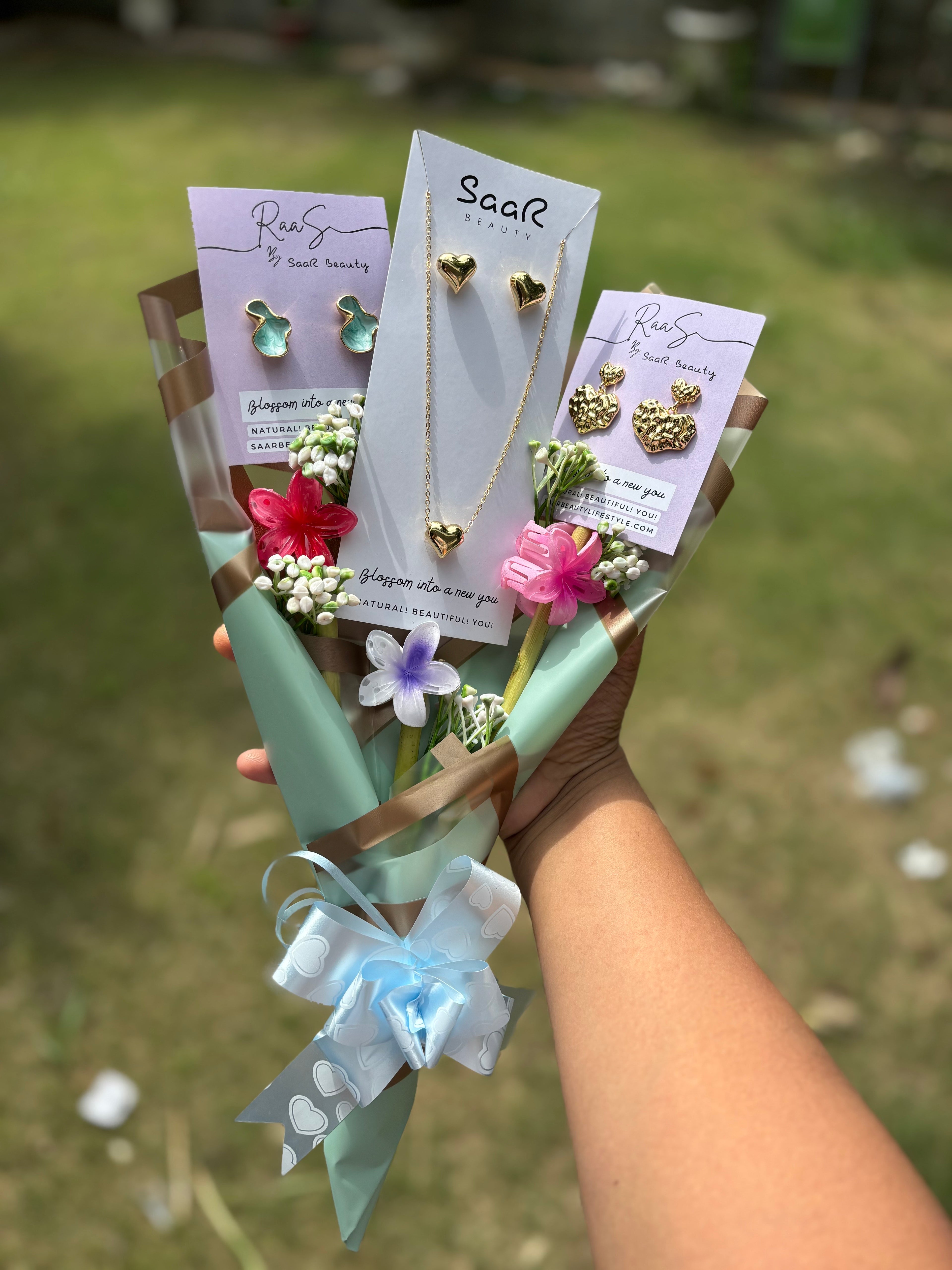 Bouquet of jewelry with cards held by a hand against a blurred green Premium gift bouquet by Saar Beauty with anti-tarnish heart necklace and earrings, pastel floral wrapping, and small flower claw hair clips. Ideal for thick hair, long hair, and gifting special women and girls.background