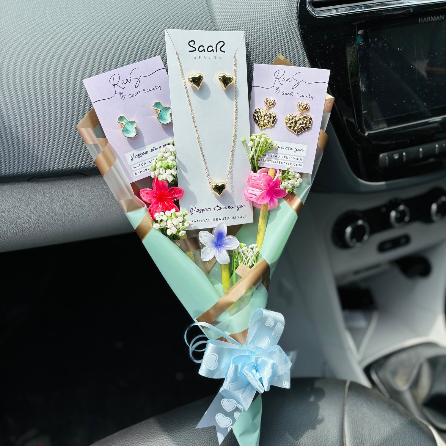 Jewelry set with necklaces and earrings displayed on a car dashboard.Premium gift bouquet by Saar Beauty with anti-tarnish heart necklace and earrings, pastel floral wrapping, and small flower claw hair clips. Ideal for thick hair, long hair, and gifting special women and girls.