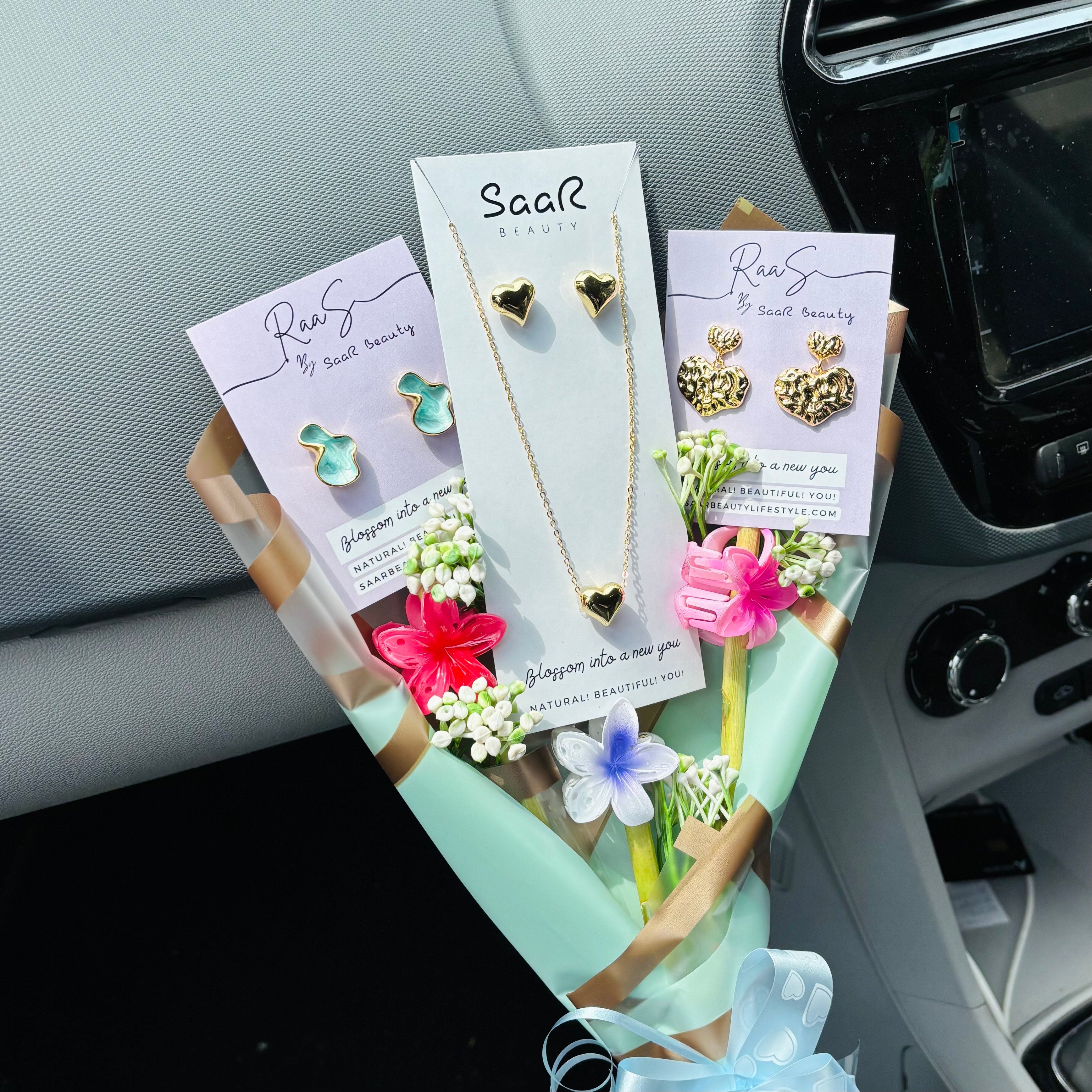 Jewelry set with necklaces and earrings on display cards, placed on a car dashboard.Premium gift bouquet by Saar Beauty with anti-tarnish heart necklace and earrings, pastel floral wrapping, and small flower claw hair clips. Ideal for thick hair, long hair, and gifting special women and girls.