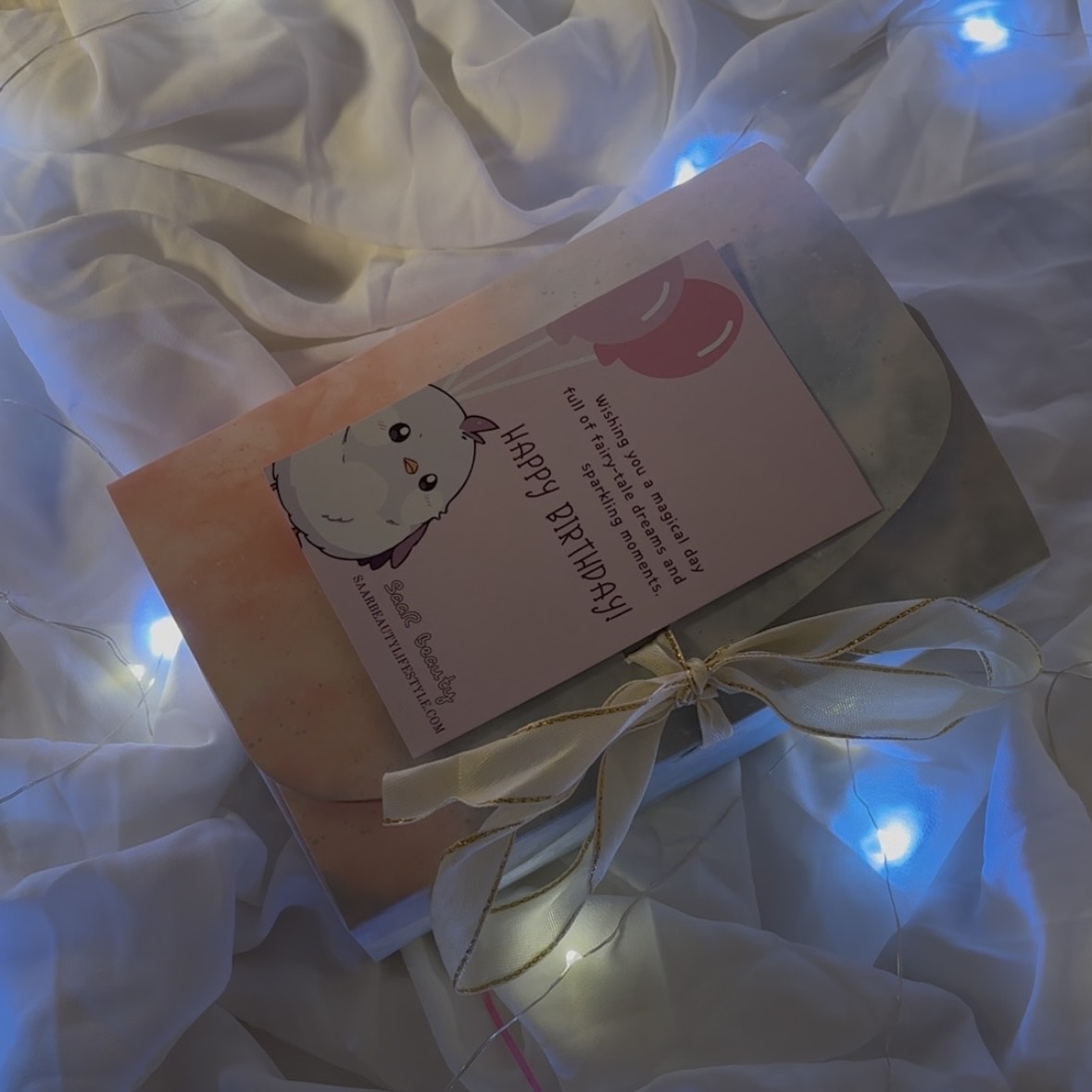 Card with a cat illustration and text on a white fabric background with string "Luxury birthday surprise gift box for girls with chocolates and accessories"lights.