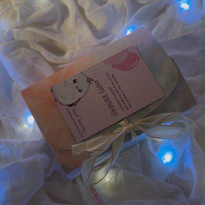 Card with a cat illustration and text on a white fabric background with string "Luxury birthday surprise gift box for girls with chocolates and accessories"lights.