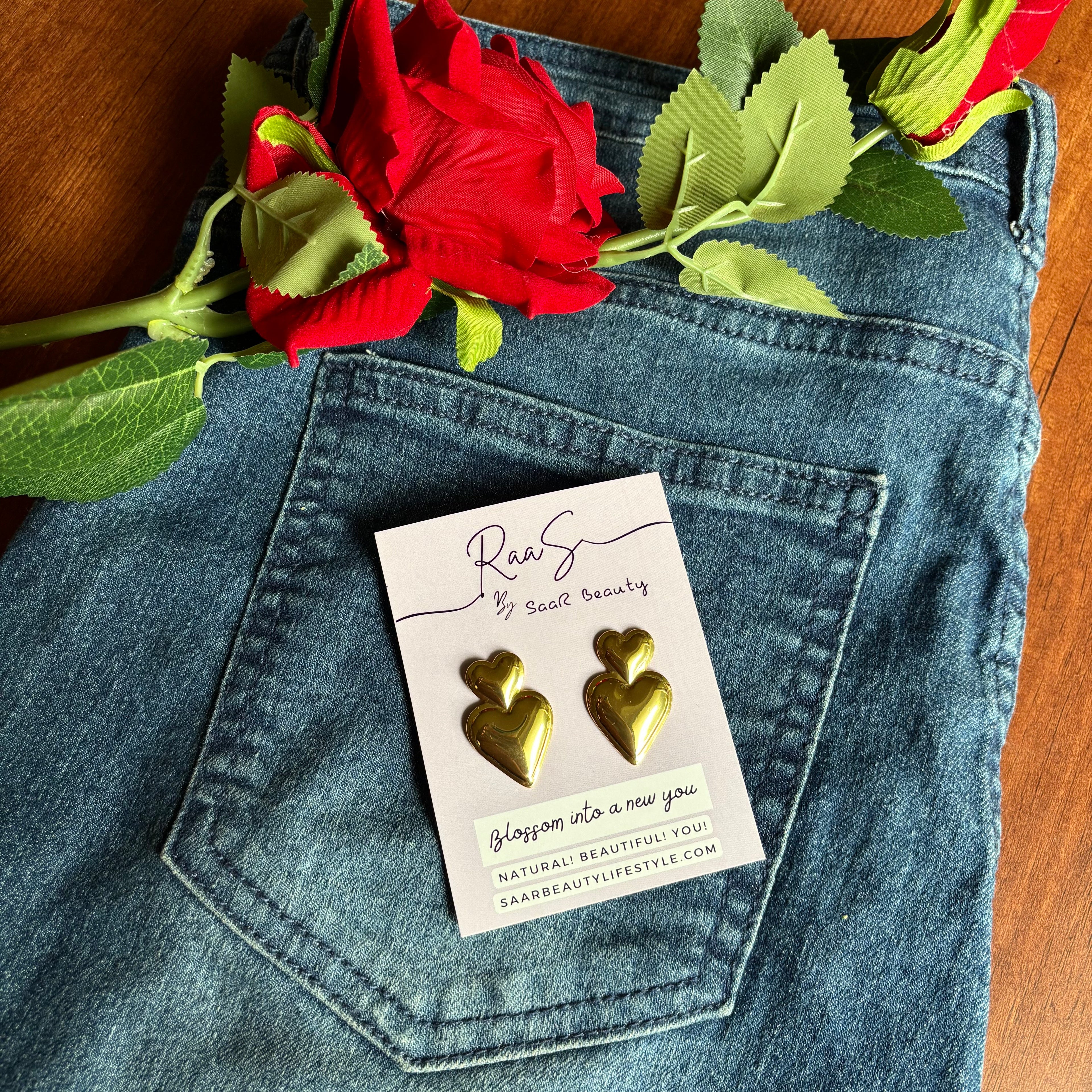 Gold heart-shaped earrings on a card with a red rose and green leaves on blue jeans. Premium anti-tarnish gold heart earrings for daily wear saar beauty