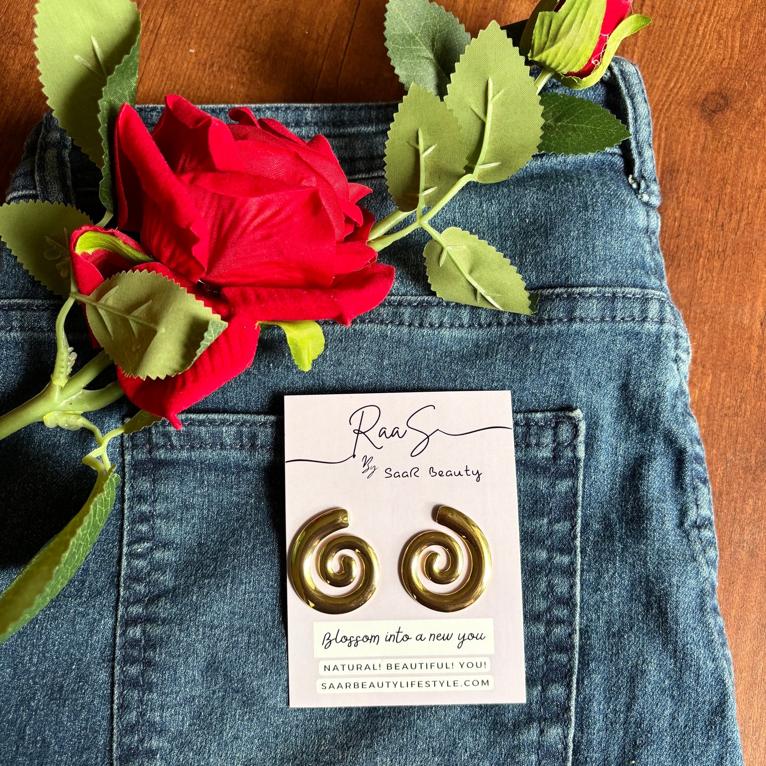 Gold spiral earrings on a card with a red rose and green leaves on blue denim fabric. 