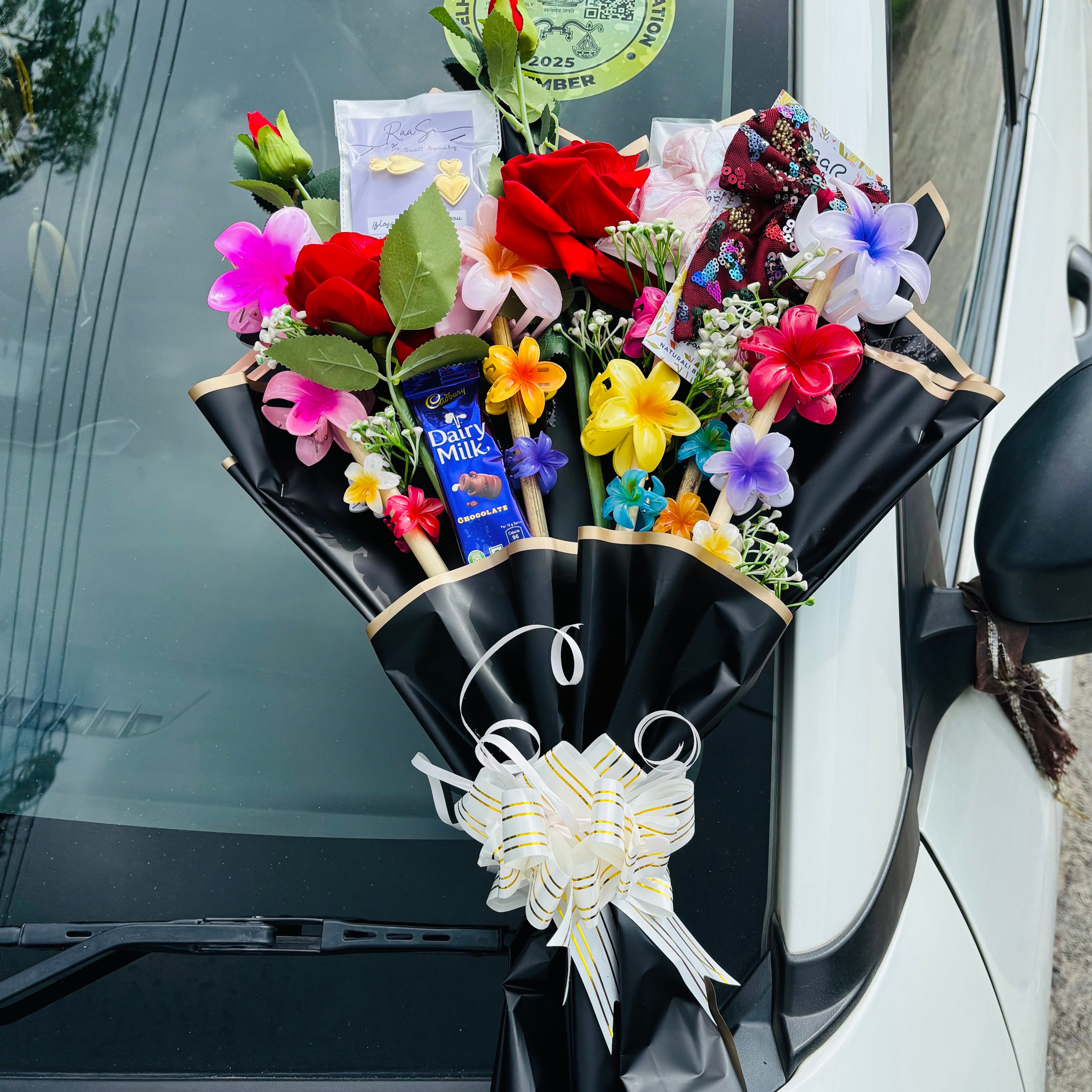 Bouquet of flowers with a white ribbon attached to a car window. Forever Love Gift Bouquet with roses, scrunchie, earrings, chocolates, hair claw clips, and bow by Saar Beauty – perfect gift for women &amp; girls