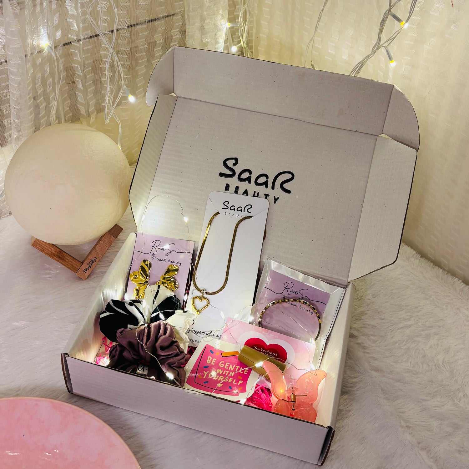 Open box from Saar Beauty with jewelry and accessories on a soft surface. Saar Beauty Buy 3 Get 5 Free Hamper – Includes waterproof jewelry, scrunchies, butterfly clip, fairy lights, and gifting card.
