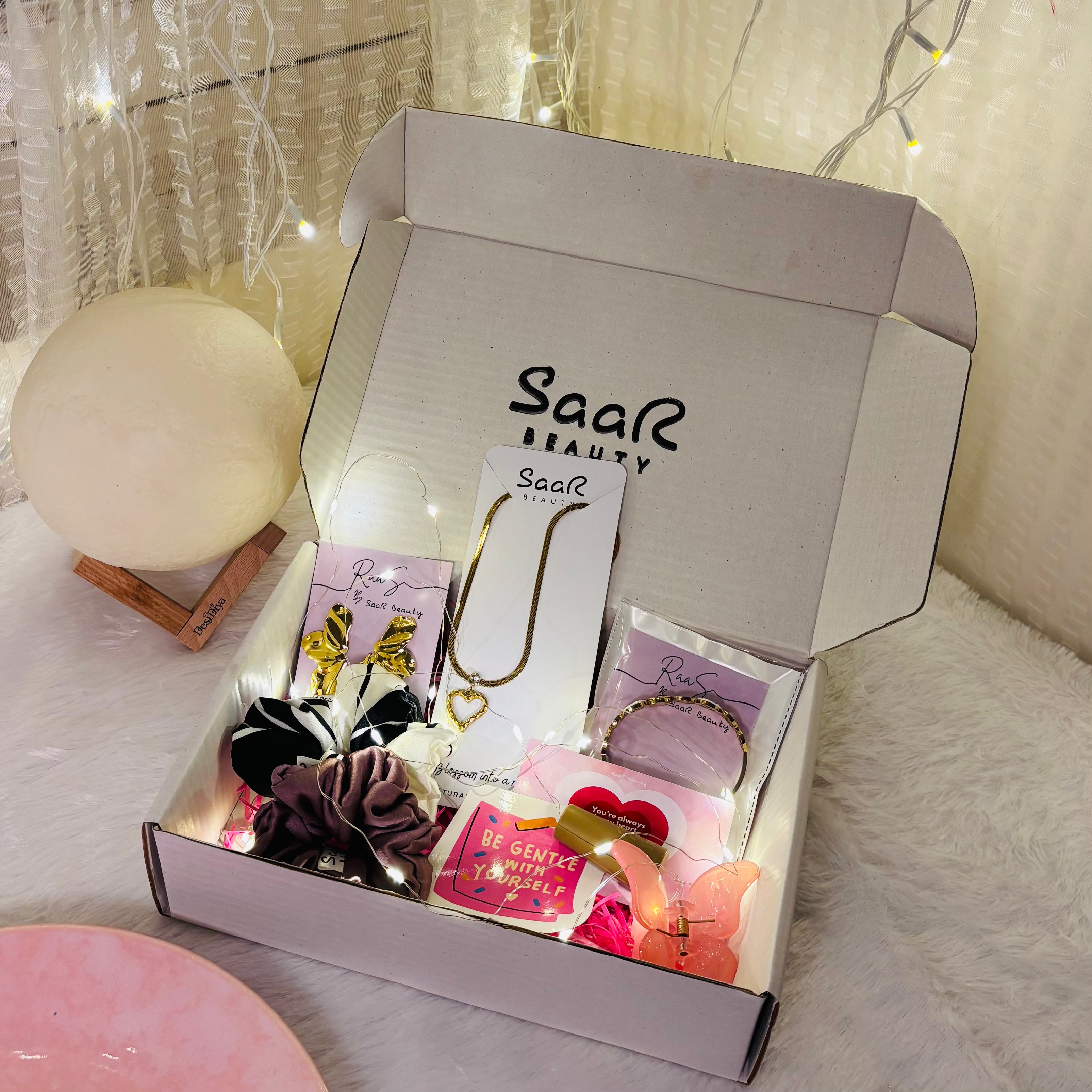 Open box from Saar Beauty with jewelry and accessories on a soft surface. Saar Beauty Buy 3 Get 5 Free Hamper – Includes waterproof jewelry, scrunchies, butterfly clip, fairy lights, and gifting card.