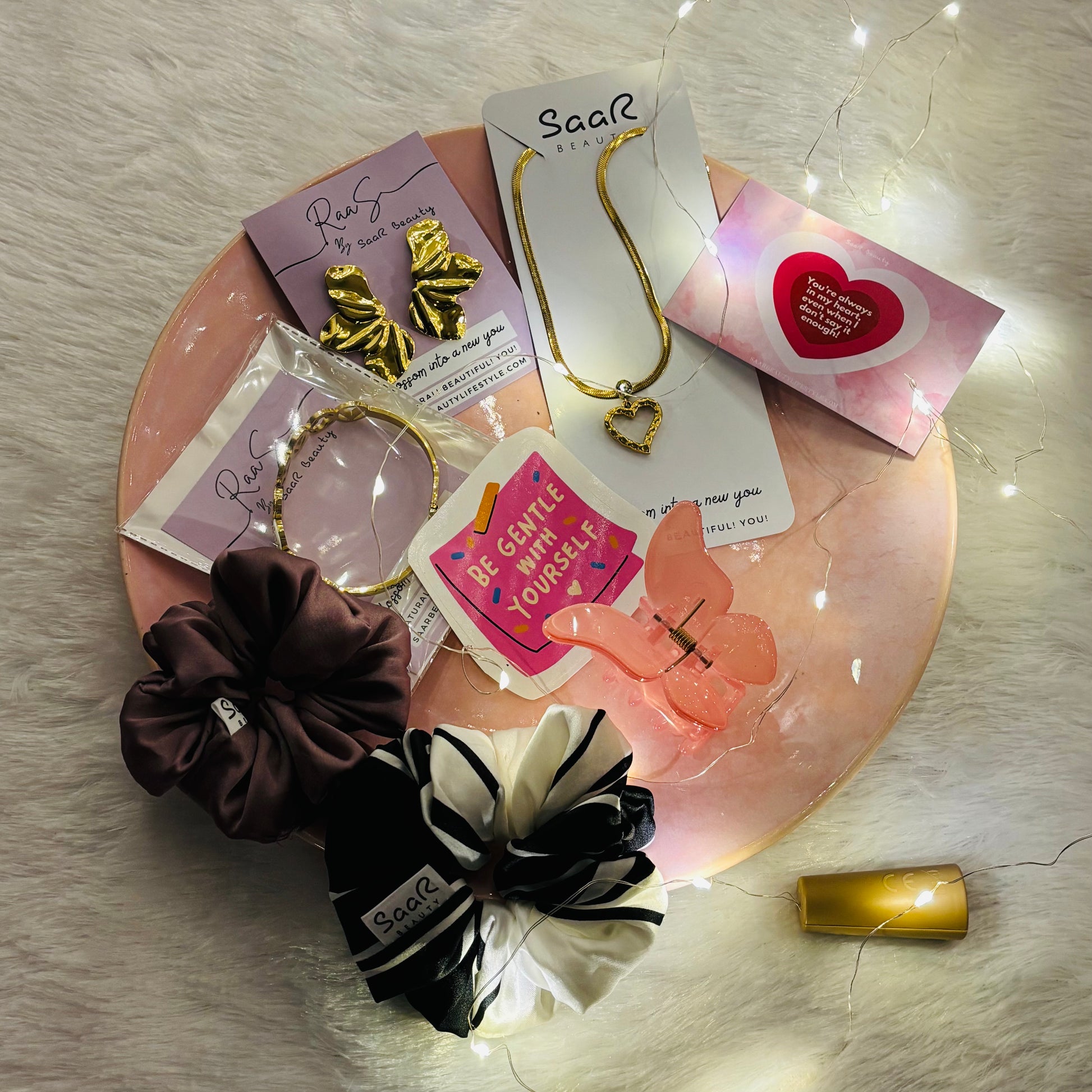 Set of hair accessories including scrunchies and a necklace on a decorative plate with fairy lights. Saar Beauty Buy 3 Get 5 Free Hamper – Includes waterproof jewelry, scrunchies, butterfly clip, fairy lights, and gifting card.