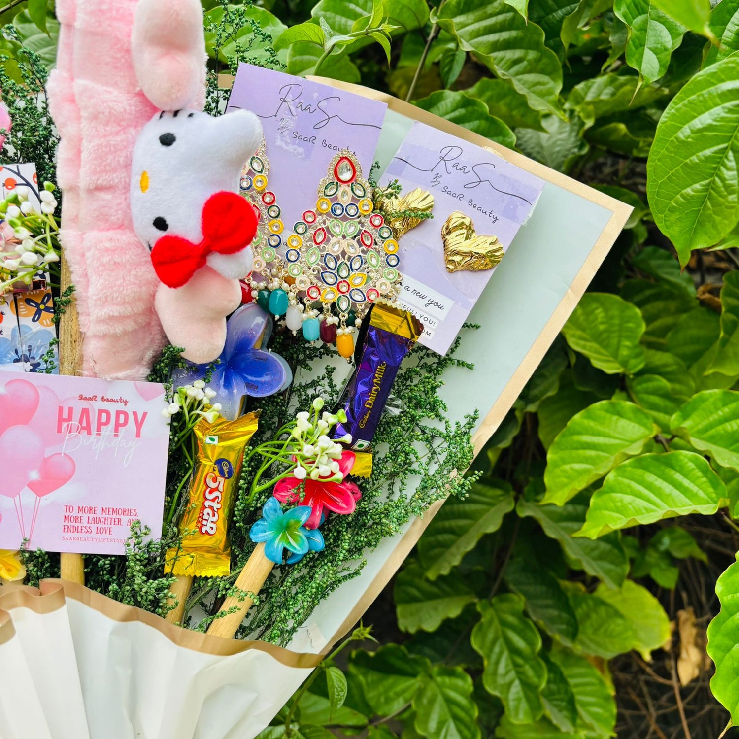 Bouquet with plush toy, candy, and cards against a green leafy background Saar Beauty Limited Edition Birthday Accessories Bouquet – unique floral gift bouquet with hair clutchers, earrings, chocolates, and accessories for women and girl