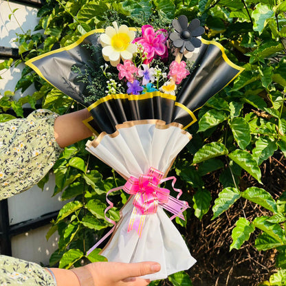 Bouquet of flowers wrapped in black paper with a pink ribbon held by a person against a green leafy background. Limited Edition Saar Beauty Hair Clutcher Bouquet – Unique floral gift with 9 colorful hair claw clips beautifully wrapped for women and girls.